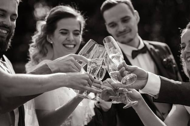 Braut und Bräutigam trinken Champagner mit besten Freunden im Freien mit aufrichtigem Lächeln und monochromem Blick