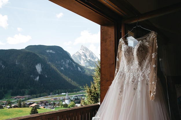 Brautkleid hängt an einem Kleiderbügel an einem Fenster mit Blick auf die Berge