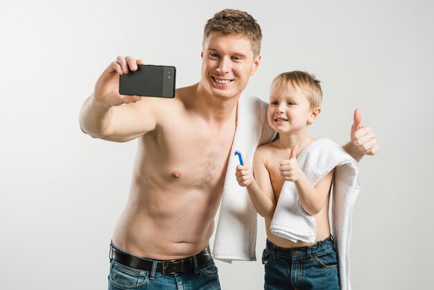 Bringen Sie und sein Sohn hervor, der Daumen herauf das Zeichen zeigt, das selfie am Handy gegen grauen Hintergrund nimmt