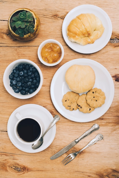 Kostenloses Foto brötchen; brot; kekse; marmelade; blaubeeren und kaffeetasse mit besteck auf hölzernen hintergrund