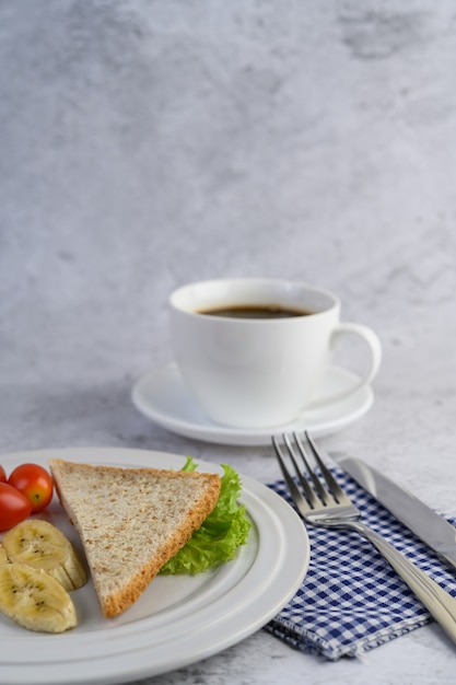 Brot, Banane und Tomate auf weißem Teller mit Kaffeetasse, Gabel und Messer.