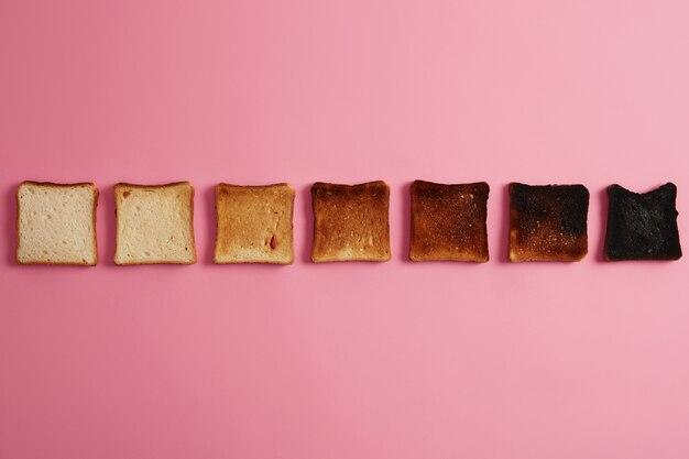 Brotscheiben in verschiedenen Röststadien. Knusprige geröstete Scheiben, angeordnet in einer Reihe über rosa Hintergrund. Letzterer ist komplett verbrannt. Toast machen. Von ungeröstet bis verkohlt. Ansicht von oben nach unten