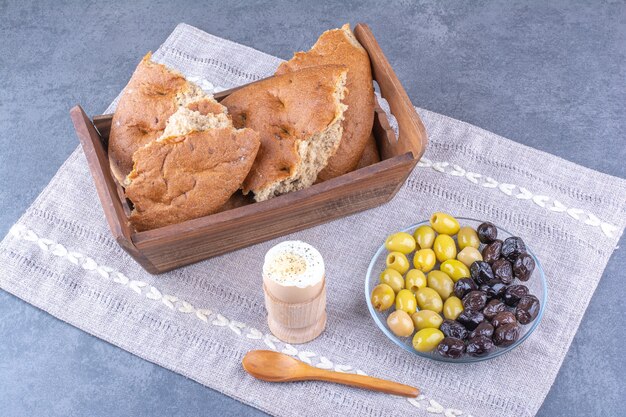 Brottablett, weich gekochtes Ei und eine Platte mit kernlosen Oliven auf einer kleinen Tischdecke auf Marmoroberfläche