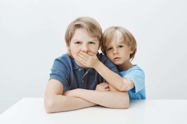 Bruder bittet darum, geheim zu bleiben. Porträt eines unglücklichen verärgerten Jungen, der mit gekreuzten Händen am Tisch sitzt und die Stirn runzelt, während Geschwister seinen Mund mit Handfläche bedecken