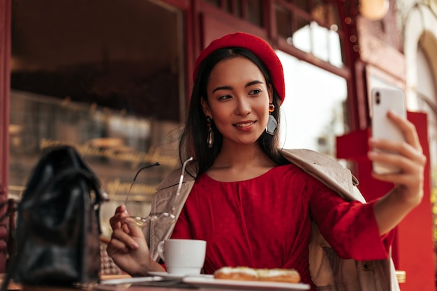 Brünette asiatische braunäugige Frau in stilvoller Baskenmütze, rotem Kleid, beigem Trenchcoat sitzt in einem gemütlichen Straßencafé, hält trendige Brillen und macht Selfies