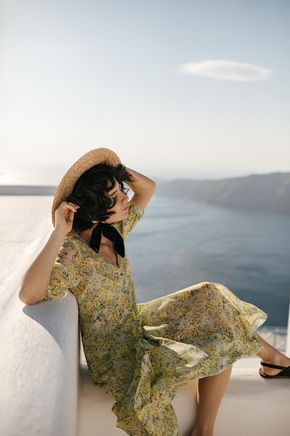Brünette attraktive Dame in geblümtem Hut und Midikleid sitzt auf der Terrasse mit Meerblick Lockige Frau im Bootsfahrer genießt die Sonne auf dem Balkon auf Meereshintergrund