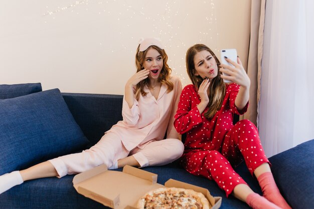 Brünette Dame, die Telefon für Selfie mit Freund benutzt und lustige Gesichter macht. Innenfoto von zwei Schwestern im niedlichen Pyjama, der Pizza zusammen isst.