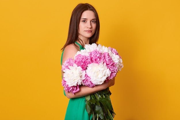 Brünette Frau, die großen Blumenstrauß mit rosa und weißen Pfingstrosen umarmt, stilvolle Frau mit Blumen, hat ruhigen Gesichtsausdruck und posiert isoliert auf Gelb.