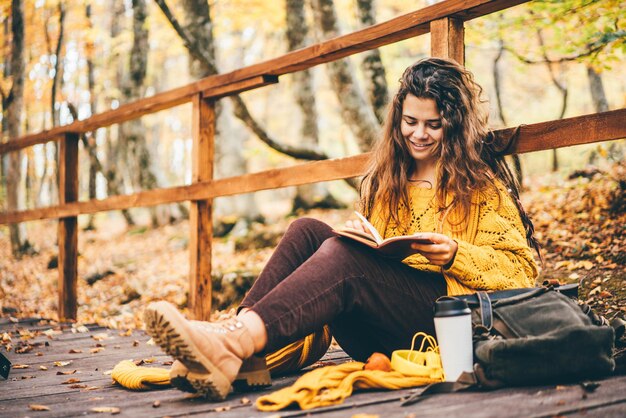 Brünette frau im gelben pullover sitzt auf einem gefallenen herbstlaub in einem park, liest ein buch oder schreibt ein tagebuch
