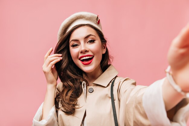 Brünette Frau in beige Baskenmütze lächelt und macht Selfie auf rosa Hintergrund.