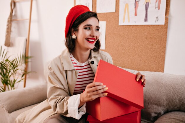 Brünette Frau in beige Graben lächelt und öffnet rote Box. Schönes reizendes Mädchen im gestreiften Pullover und im hellen Hut lächelt.