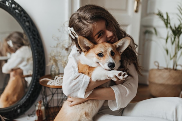 Brünette Frau lächelt und umarmt Corgi Fröhliche junge lockige Dame in weißer Bluse hat Spaß mit süßem Hund im gemütlichen Zimmer