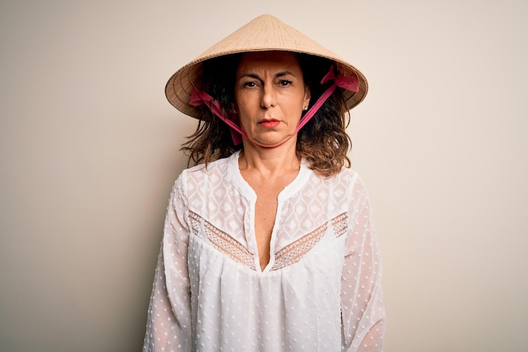 Kostenlose Foto brünette frau mittleren alters mit traditionellem asiatischen konischen hut vor weißem hintergrund, skeptisch und nervös, runzelt die stirn, verärgert über das problem einer negativen person