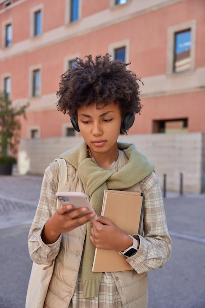 Brünette Frau studiert online hört pädagogischen Vortrag mit Smartphone und Kopfhörer gehen im Freien spazieren und tragen Notizblöcke in Freizeitkleidung.