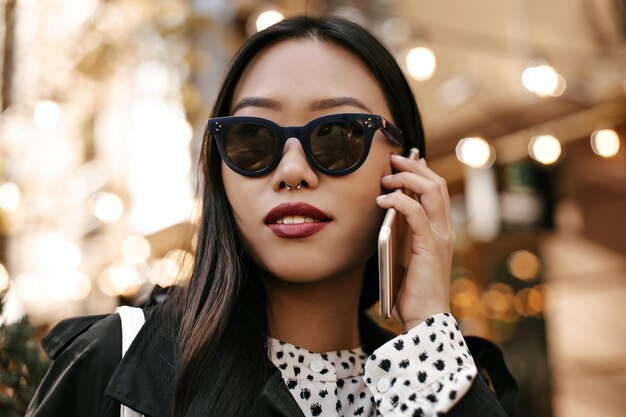 Brünette gebräunte Dame mit Sonnenbrille spricht am Telefon Charmante asiatische Frau in Schwarz-Weiß-Outfit posiert draußen