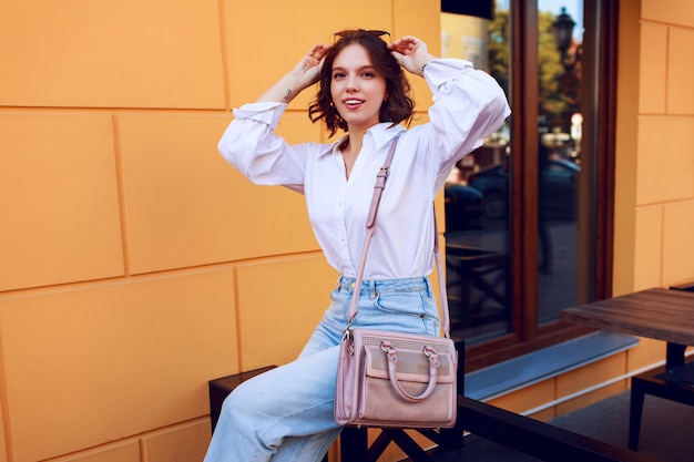 Brünette glückliche Frau mit den kurzen Haaren, die draußen sitzen. Trägt stilvolle weiße Bluse und Jeans. Modischer Herbstlook.