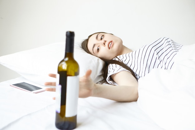 Brünette junge Frau, die morgens unter schrecklichen Kopfschmerzen leidet, im Bett liegt und nach einer Flasche Alkohol greift