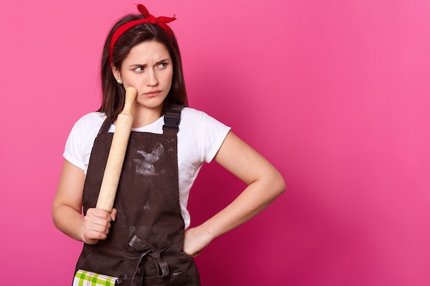 Brünette Köchin mit rotem Haarband, mit Mehl verschmutzter brauner Schürze und weißem T-Shirt entscheidet, welches Rezept zum Backen von Kuchen verwendet wird. Junger Bäcker hält Nudelholz und berührt damit die Wange. Kulinarisches Konzept.