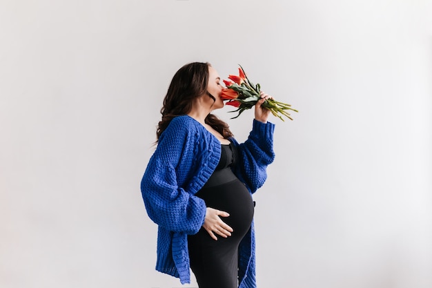 Brünette lockige Frau im schwarzen Kleid und in der blauen Strickjacke hält Tulpenstrauß. Attraktive schwangere Dame riecht Tulpen auf lokalisiertem Hintergrund.