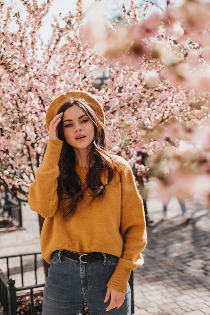 Brünette Mädchen in stilvollem Outfit und Hut posiert in der Nähe von Sakura. Porträt der Frau im orangefarbenen Pullover, in der Jeans und in der Baskenmütze, die im blühenden Garten gehen