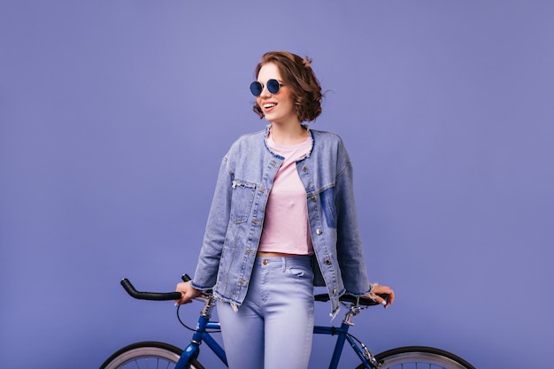 Brünette optimistische Frau in der dunklen Sonnenbrille, die mit Fahrrad aufwirft. Aktives fröhliches Mädchen in Jeanskleidung stehend.