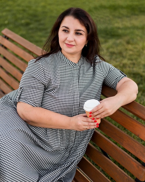 Brünette Smiley-Frau, die eine Tasse Kaffee hält, während sie auf einer Bank sitzt