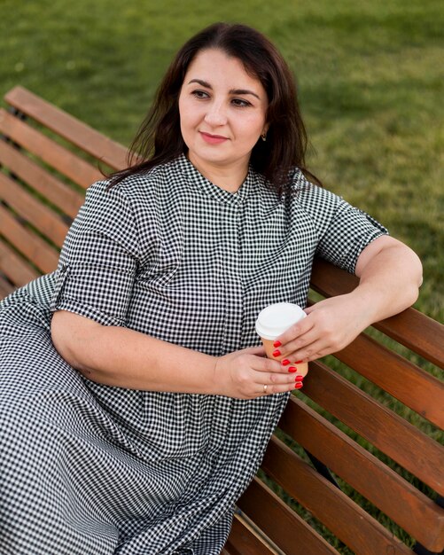 Brünette Smiley-Frau, die eine Tasse Kaffee hält, während sie auf einer Bank sitzt
