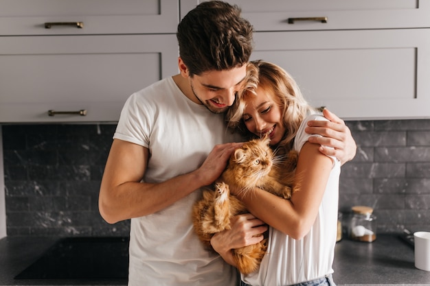 Brünetter Mann, der seine Katze betrachtet und Frau umarmt. Innenporträt der glücklichen Familie, die mit Haustier aufwirft.