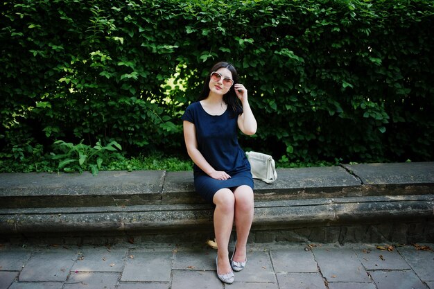 Brünettes Geschäftsmädchen im schwarzen Kleid mit Sonnenbrille sitzt an der Grenze gegen Büsche und posiert auf der Straße der Stadt