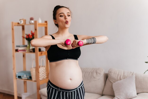 Brünettes Mädchen atmet tief durch und macht Sportübungen. Schwangere Frau im schwarzen Oberteil hält rosa Hanteln im Wohnzimmer.