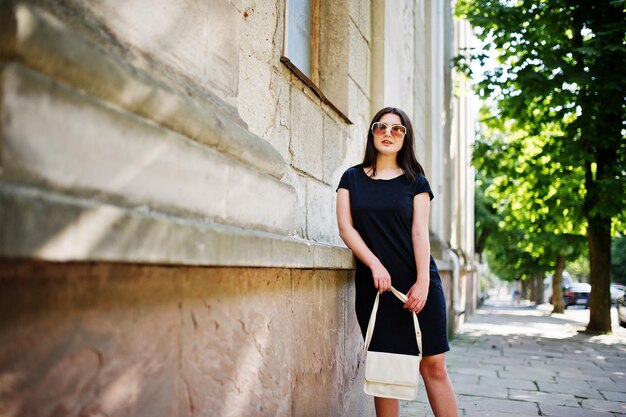 Brünettes Mädchen im schwarzen Kleid auf Sonnenbrille mit Handtasche zur Hand posiert auf der Straße der Stadt