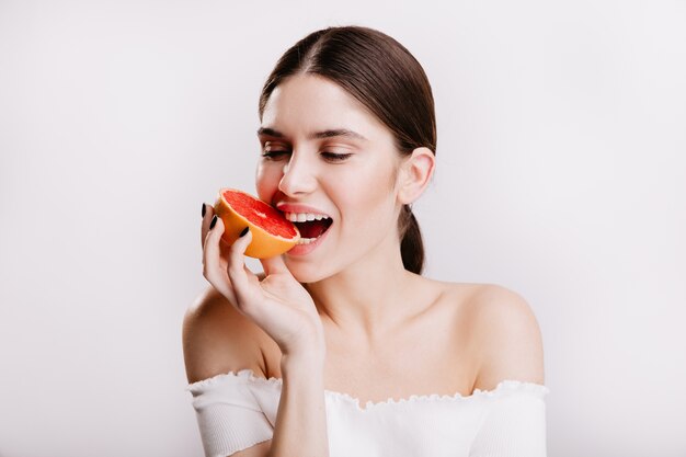 Brünettes Mädchen mit gesunder Haut beißt saftige rote Grapefruit. Nahaufnahmeporträt der Frau in großer Stimmung auf isolierter Wand.