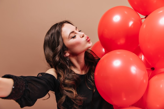 Brünettes Mädchen mit welligem Haar nimmt Selfie, bläst Kuss und hält rote Luftballons.