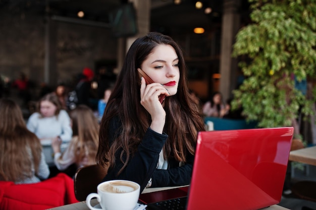 Brünettes Mädchen sitzt im Café, arbeitet mit rotem Laptop und spricht am Handy