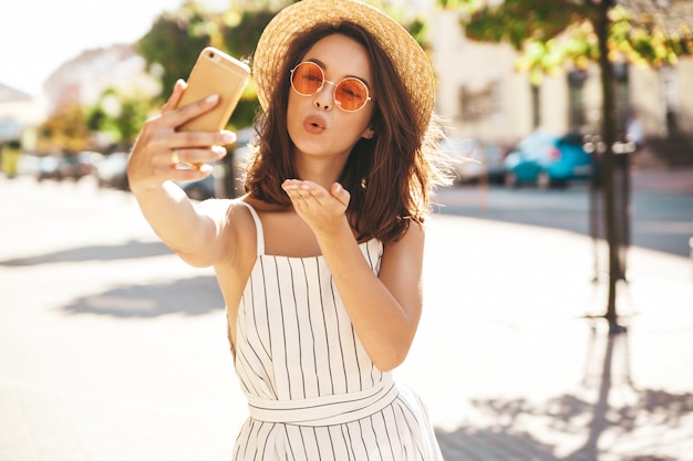 Brünettes modell in sommerkleidung, die auf der straße unter verwendung des mobiltelefons posiert, das luftkuss gibt