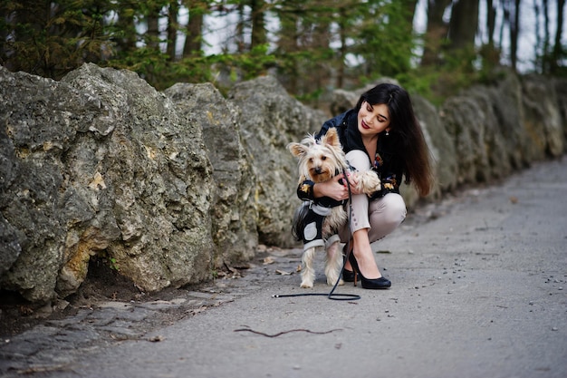 Brünettes Zigeunermädchen mit Yorkshire-Terrier-Hund posierte gegen Steine im Park Model Wear auf Lederjacke und T-Shirt mit Zierhosen und Schuhen mit hohen Absätzen