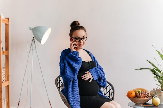 Kostenloses Foto brunette schwangere frau in brillen und blaue strickjacke am telefon sprechen charmante dame in schwarzem kleid posiert im wohnzimmer