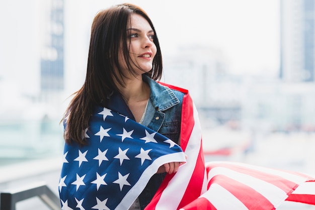 Brunettefrau eingewickelt in der amerikanischen Flagge auf Hintergrund der Stadt