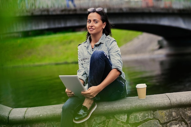 Brunettefrau sitzt auf einer Brücke und hält Tablet-PC.