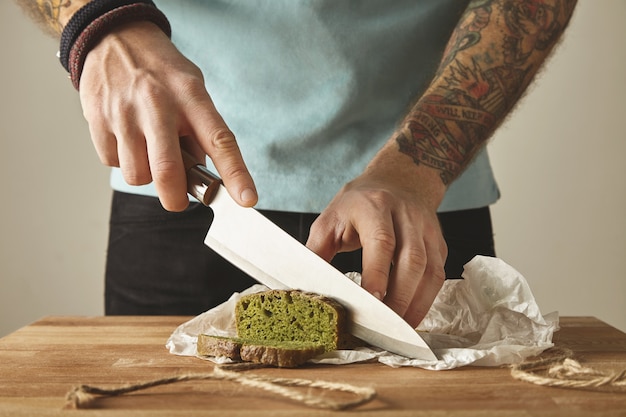 Brutal Mann tätowierte Hände schneiden gesunde Spinat hausgemachte grüne rustikale Brot mit Vintage-Messer auf Scheiben. Weißer Tisch aus Holzbrett