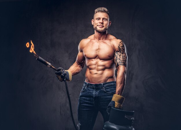 Brutal tätowierter männlicher Schweißer mit stylischem Haarschnitt und Bart, mit muskulösem Körper, nur mit Jeans bekleidet, hält einen Propantank und einen brennenden Brenner, steht in einem Studio und schaut mit einem Grinsen. Isoliert an