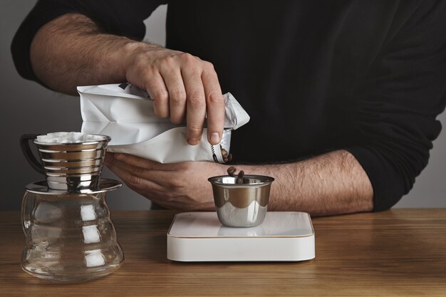 Brutaler Barista in schwarzem Sweatshot gießt geröstete Kaffeebohnen in eine rostfreie Tasse. Chrom-Filterkaffeemaschine in der Nähe im Café-Laden