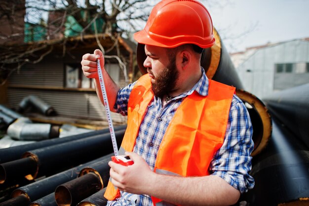 Brutaler Bart Arbeiter Mann Anzug Bauarbeiter in orangefarbenem Sicherheitshelm in der Nähe von Stahlrohren mit Maßband zur Hand