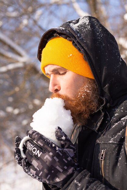 Brutaler erwachsener Mann in einem orange hellen Hut in einem verschneiten Wald an einem sonnigen Tag mit einer Tasse gefüllt mit Schnee, der Spaß hat