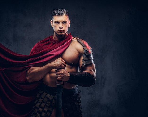 Brutaler Krieger aus dem antiken Griechenland mit einem muskulösen Körper in Kampfuniformen, der auf dunklem Hintergrund posiert.