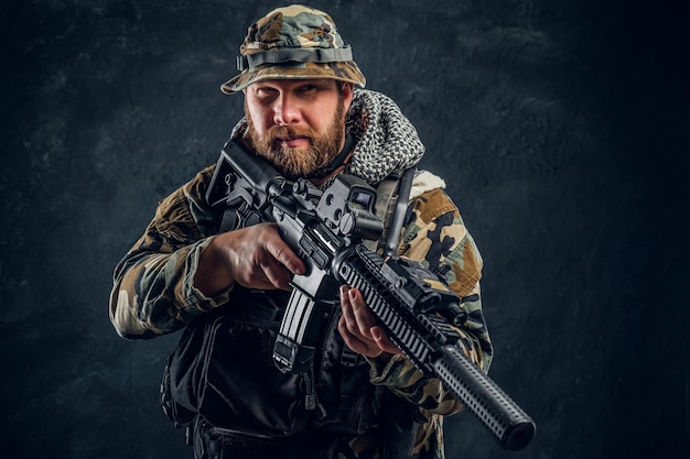 Brutaler Mann in der getarnten Militäruniform, die ein Sturmgewehr hält. Studiofoto gegen eine dunkle strukturierte Wand
