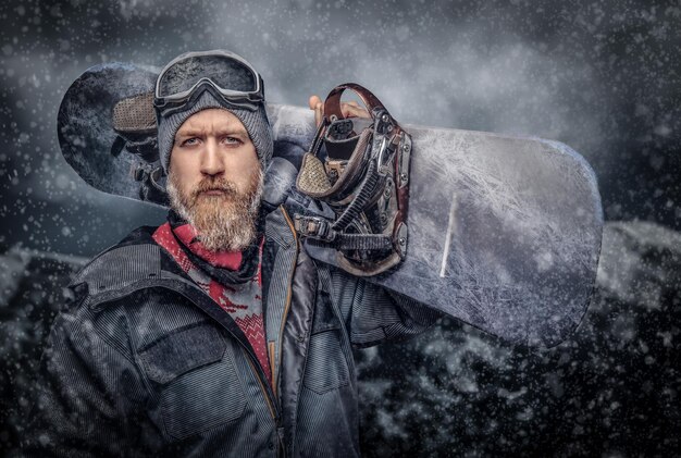 Brutaler rothaariger Snowboarder mit Vollbart in Wintermütze und Schutzbrille in einem Snowboardmantel, der mit einem Snowboard vor dem Hintergrund der Berge posiert und in eine Kamera blickt.