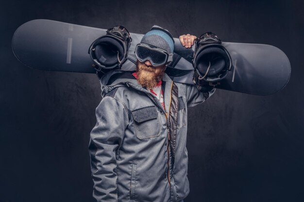 Brutaler rothaariger Snowboarder mit Vollbart in Wintermütze und Schutzbrille in Snowboardmantel hält in einem Studio ein Snowboard auf der Schulter. Isoliert auf grauem Hintergrund.