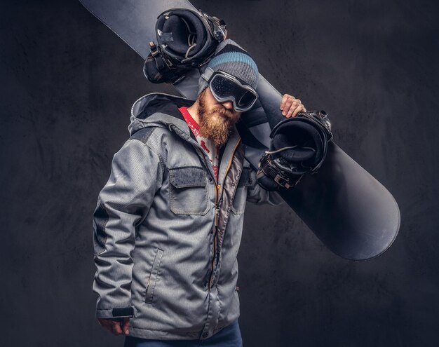 Brutaler rothaariger Snowboarder mit Vollbart in Wintermütze und Schutzbrille in Snowboardmantel hält in einem Studio ein Snowboard auf der Schulter. Isoliert auf grauem Hintergrund.