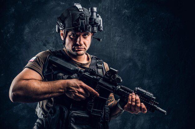 Brutaler Soldat der Russischen Föderation mit Körperschutz und Helm mit Nachtsicht, der mit einem Sturmgewehr posiert. Studiofoto gegen eine dunkle strukturierte Wand